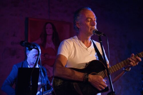 Un chanteur qui chante devant un micro avec sa guitare noire et en arrière plan un autre guitariste